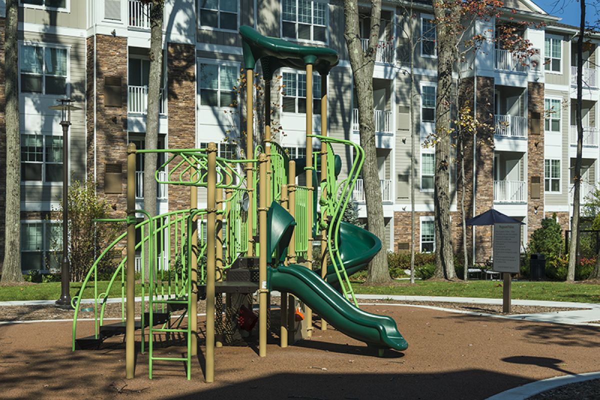 Green playground structure with slides in front of modern apartment buildings on a sunny day.