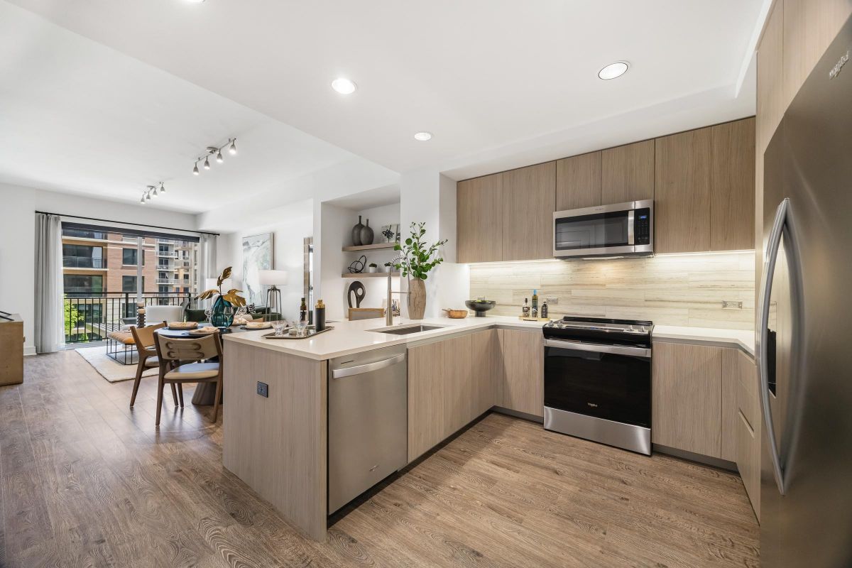 Modern kitchen with wood cabinets, stainless steel appliances, a dining area, and a living room with large windows.
