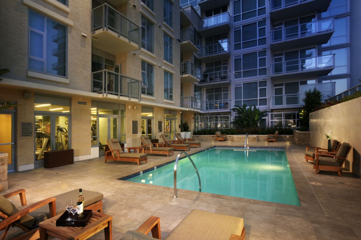 Modern apartment courtyard with a lit swimming pool, lounge chairs, and tables at dusk.