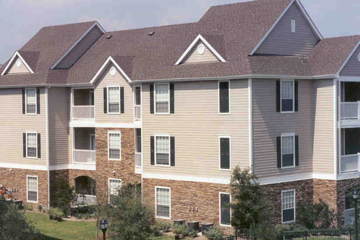 Three-story apartment building with beige siding, brick accents, balconies, and multiple windows.