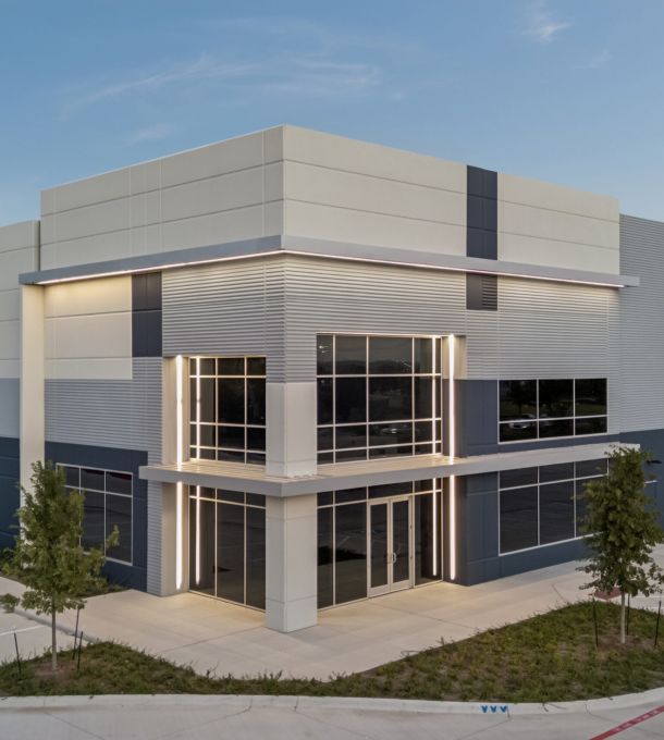 Modern industrial warehouse building with large windows, surrounded by pavement and a few small trees.