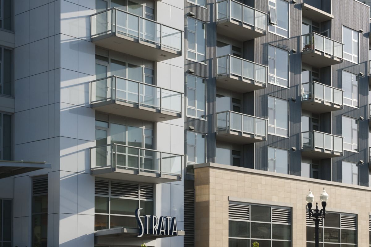 Modern apartment building with glass balconies and "Strata" signage at the entrance on a sunny day.
