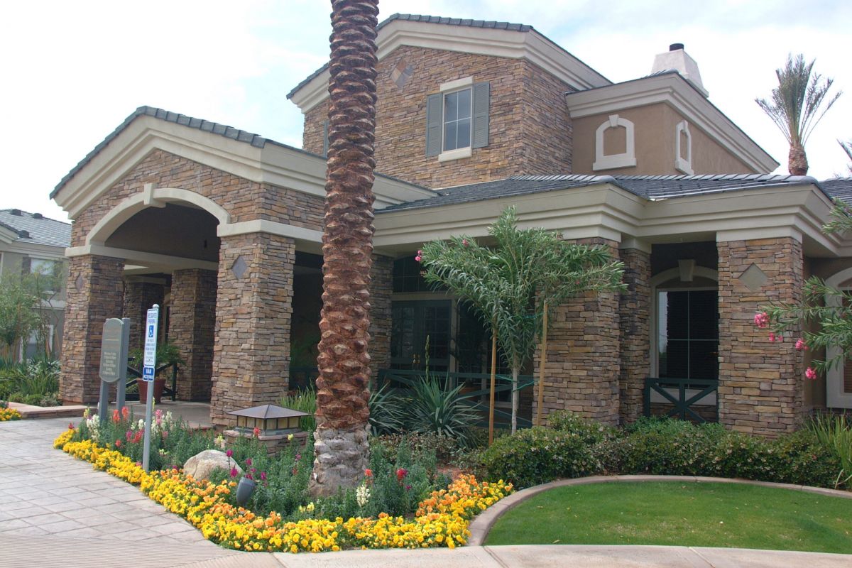 A stone-fronted building with large pillars, palm trees, and colorful flowers in the landscaped garden.