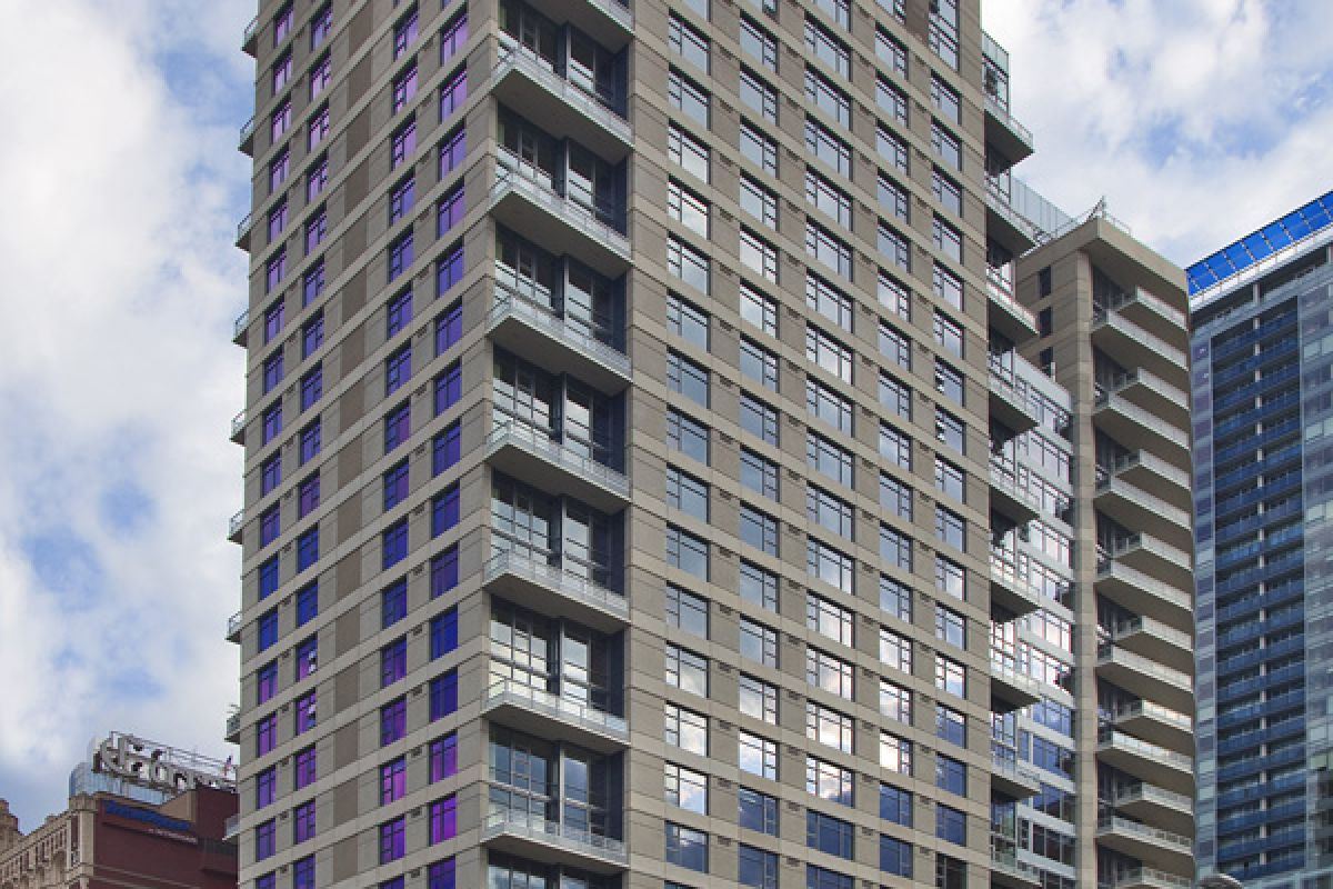 Tall modern apartment building with balconies, set against a partly cloudy sky and neighboring city buildings.