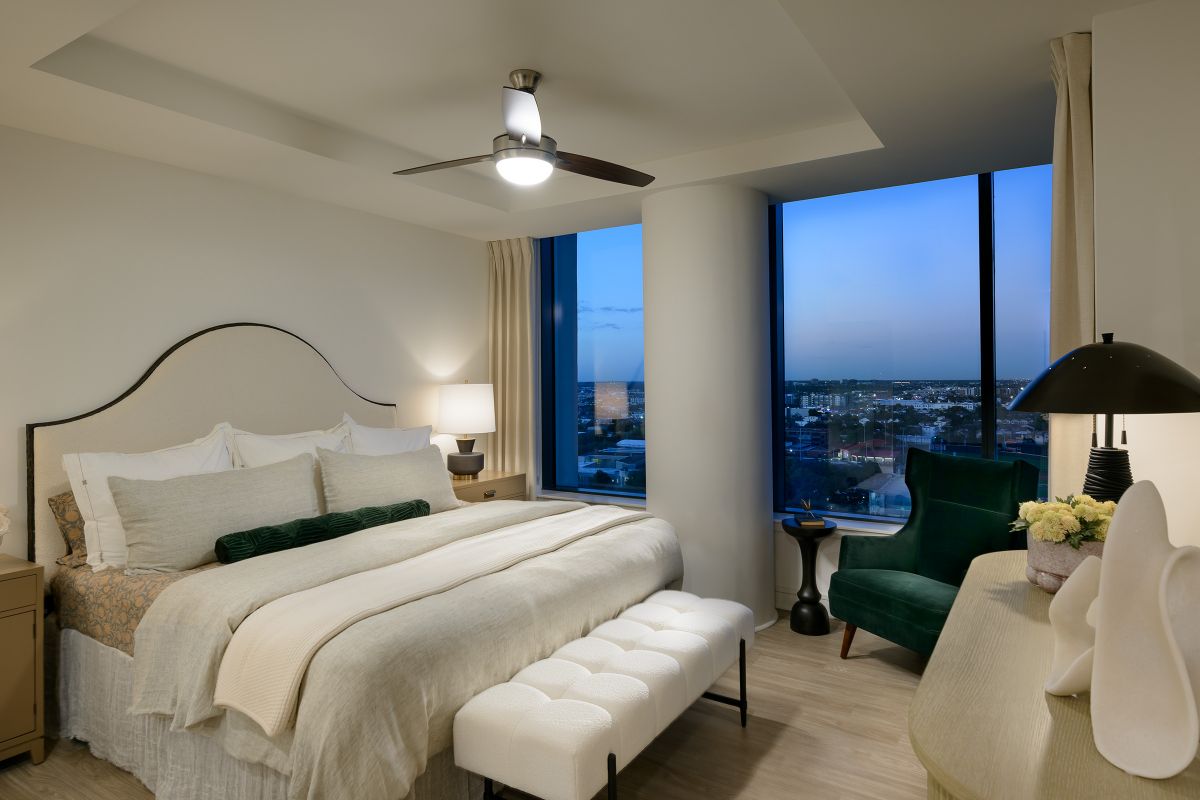 Modern bedroom at Hanover with a large bed, armchair, and city view overlooking Buffalo Bayou through floor-to-ceiling windows at dusk.