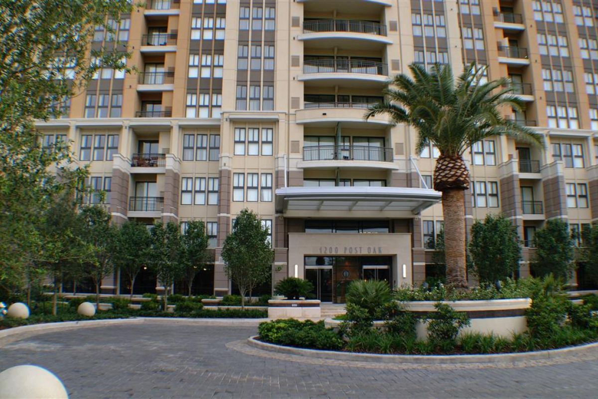 Tall modern apartment building, 1200 Post Oak, with balconies, palm tree, and a landscaped entrance roundabout.