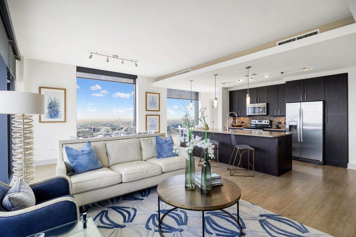 Modern living room with cream sofa, blue accents, open kitchen, and city view through large windows.