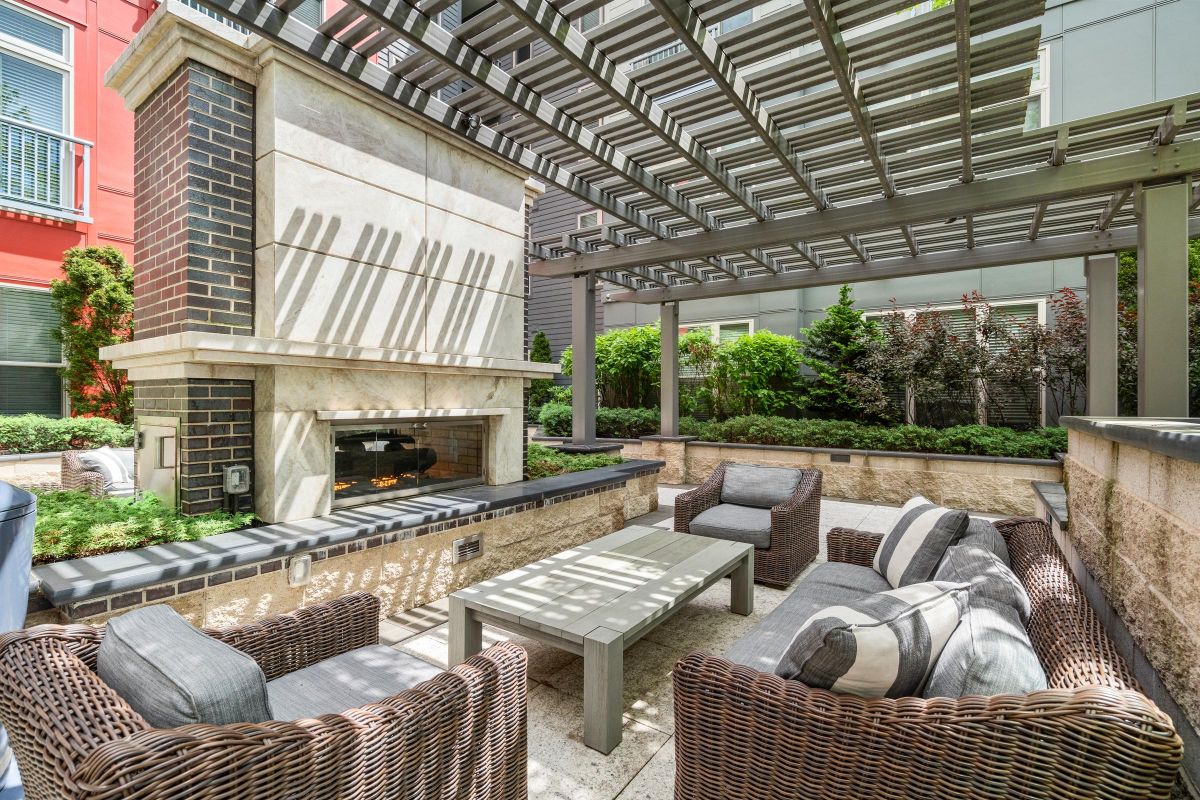 Outdoor patio with wicker sofas, a coffee table, a brick fireplace, and a pergola overhead. Lush greenery surrounds.
