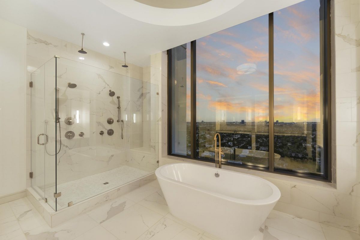 Luxurious bathroom with a glass shower, freestanding tub, and large window view of a sunset sky.