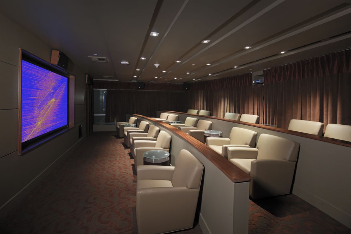 Small private movie theater with beige seats, brown carpet, and a large screen displaying abstract colors.