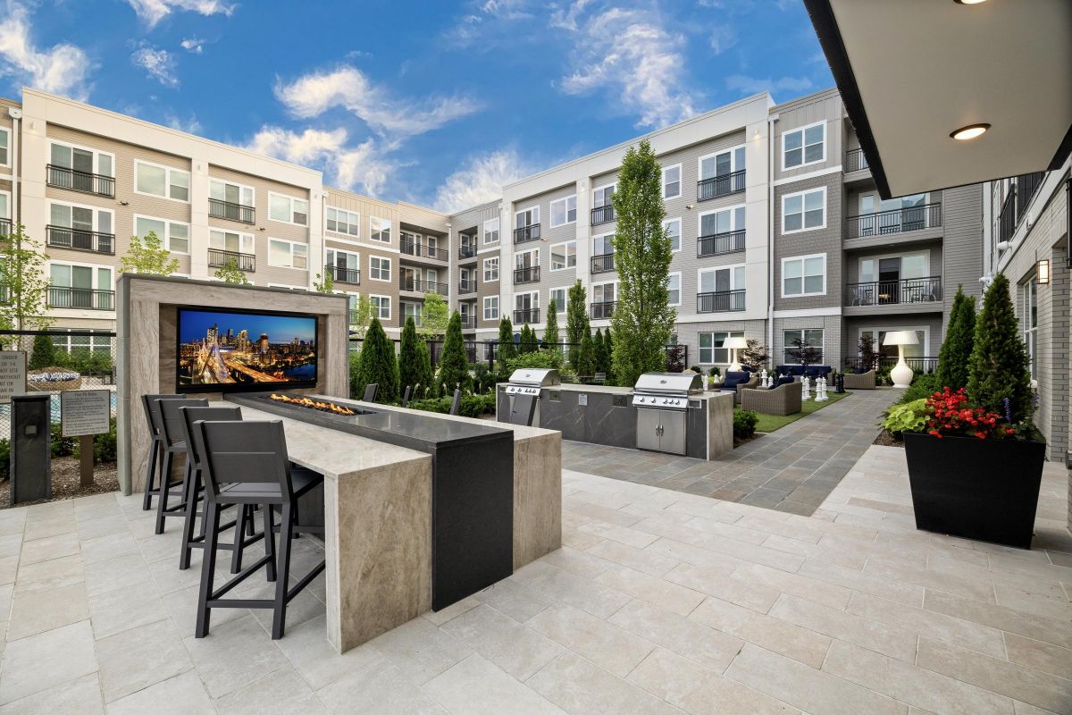 Modern apartment courtyard with seating area, fire pit, grills, potted plants, and a TV under a clear blue sky at Hanover Pinehills.