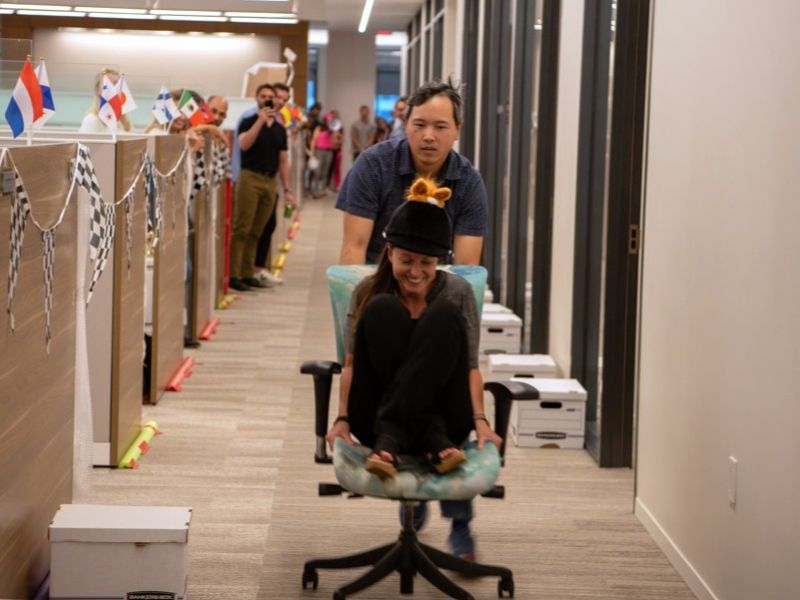 A man pushes a woman on a rolling office chair during a fun race in a decorated office hallway.