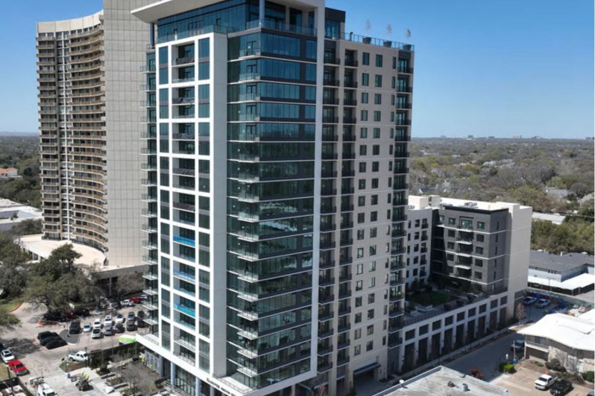 Tall modern apartment building with glass windows, surrounded by trees and other buildings under a clear blue sky.