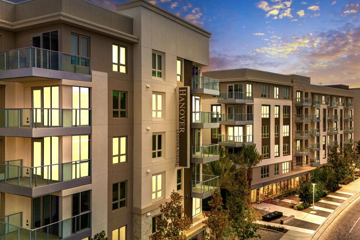 Hanover Laguna Niguel Modern apartment buildings at dusk with illuminated windows and a street lined with trees.