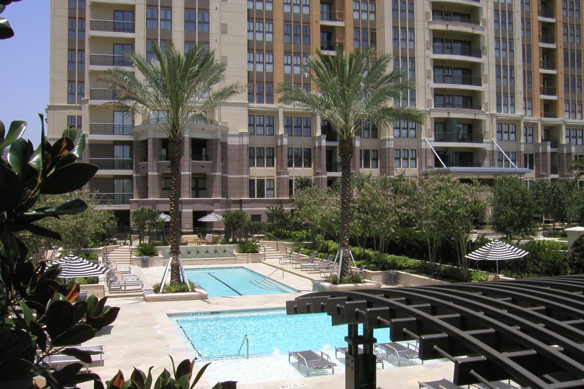 1200 Post Oak features a tall apartment building, palm trees, and a swimming pool surrounded by lounge chairs and lush greenery.