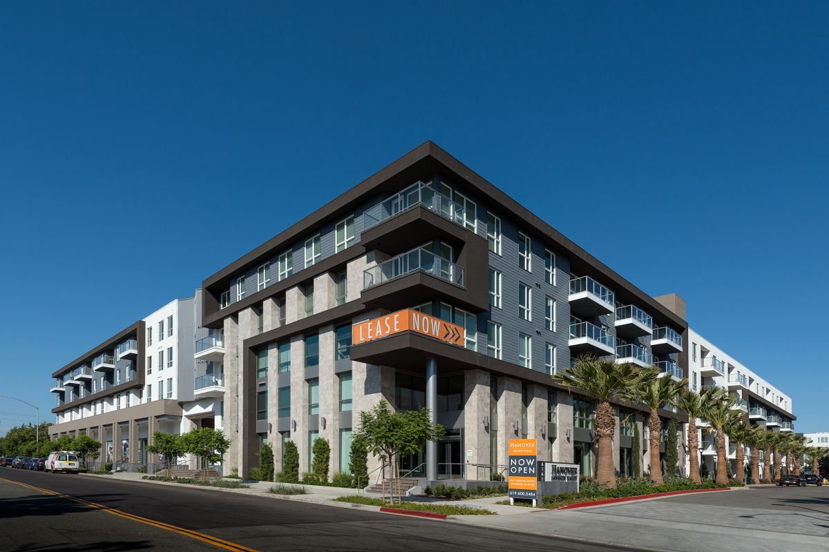 Modern apartment building with "Lease Now" and "Now Open" signs, palm trees, and clear blue sky.