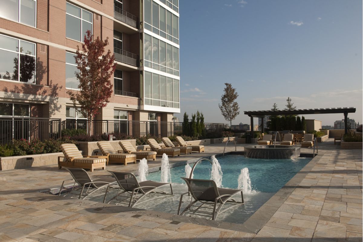 Modern apartment pool area with lounge chairs, water fountains, and a shaded pergola under a clear sky.