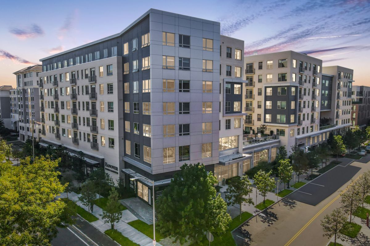 Modern multi-story apartment buildings at sunset with trees lining the street and sidewalks in the foreground.