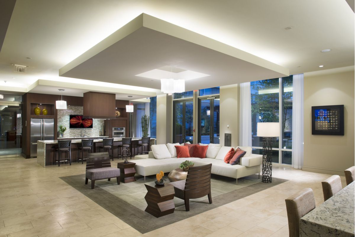 Open-plan modern living room with white sectional sofa, kitchen in background, and large windows.