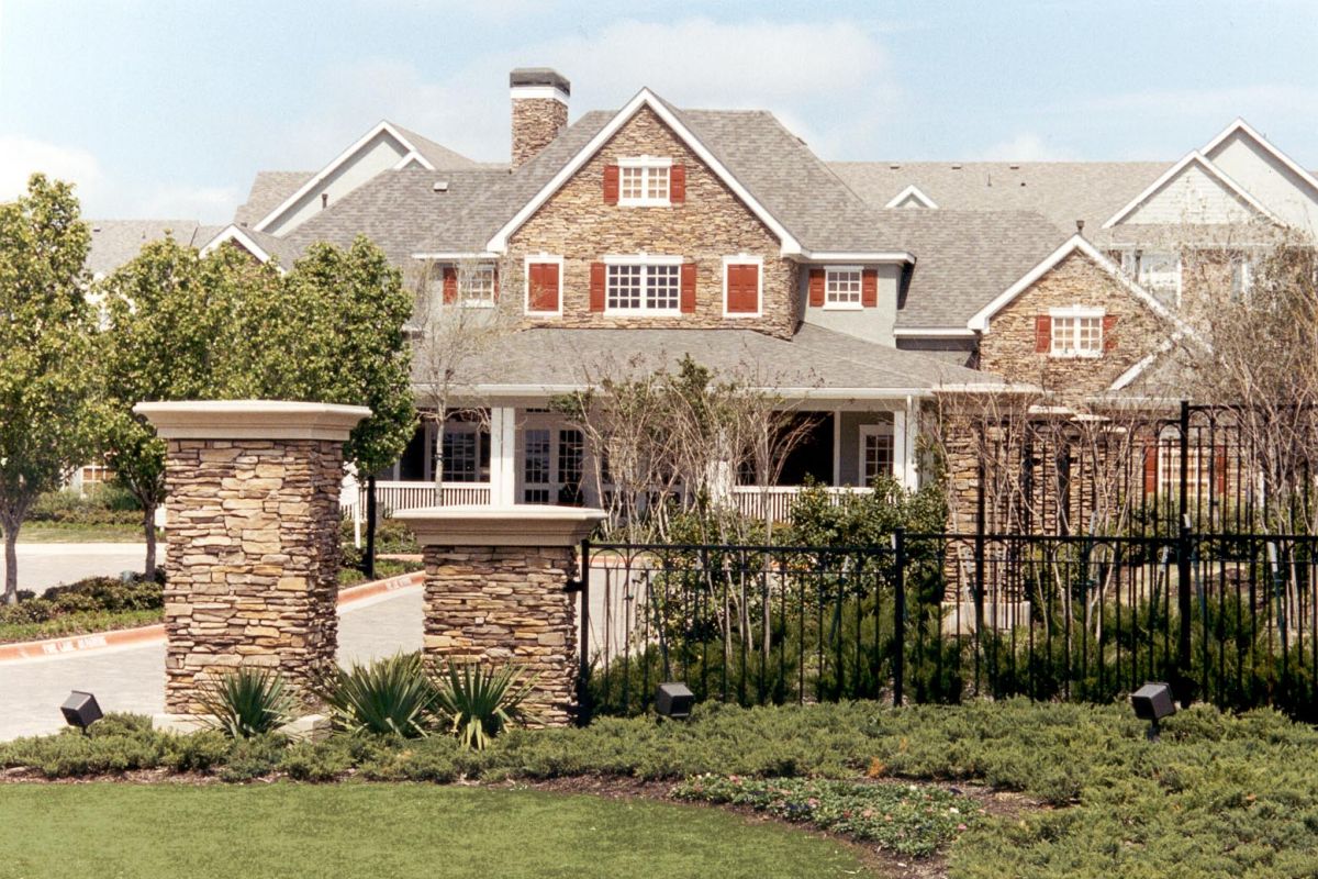 Large stone house with red shutters, front porch, and manicured lawn, behind a black metal fence and stone pillars.