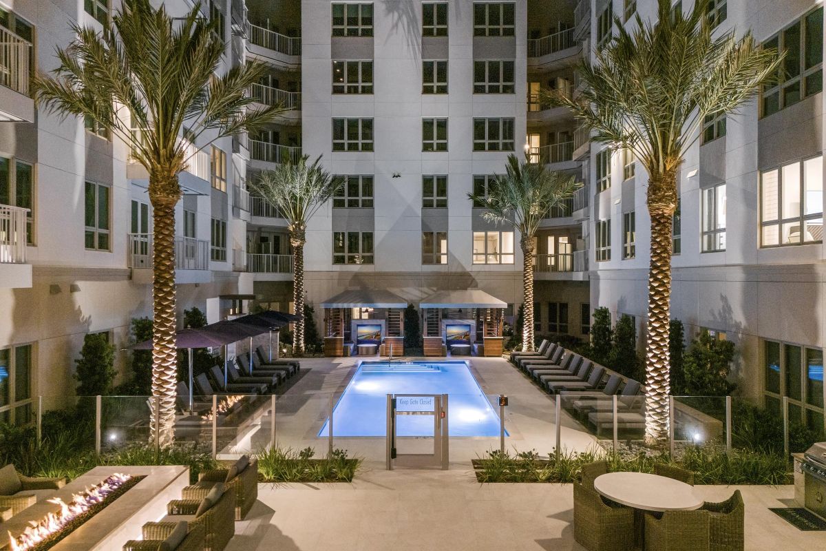 Night view of a modern apartment courtyard with a lit pool, surrounded by palm trees, seating, and a fire pit.