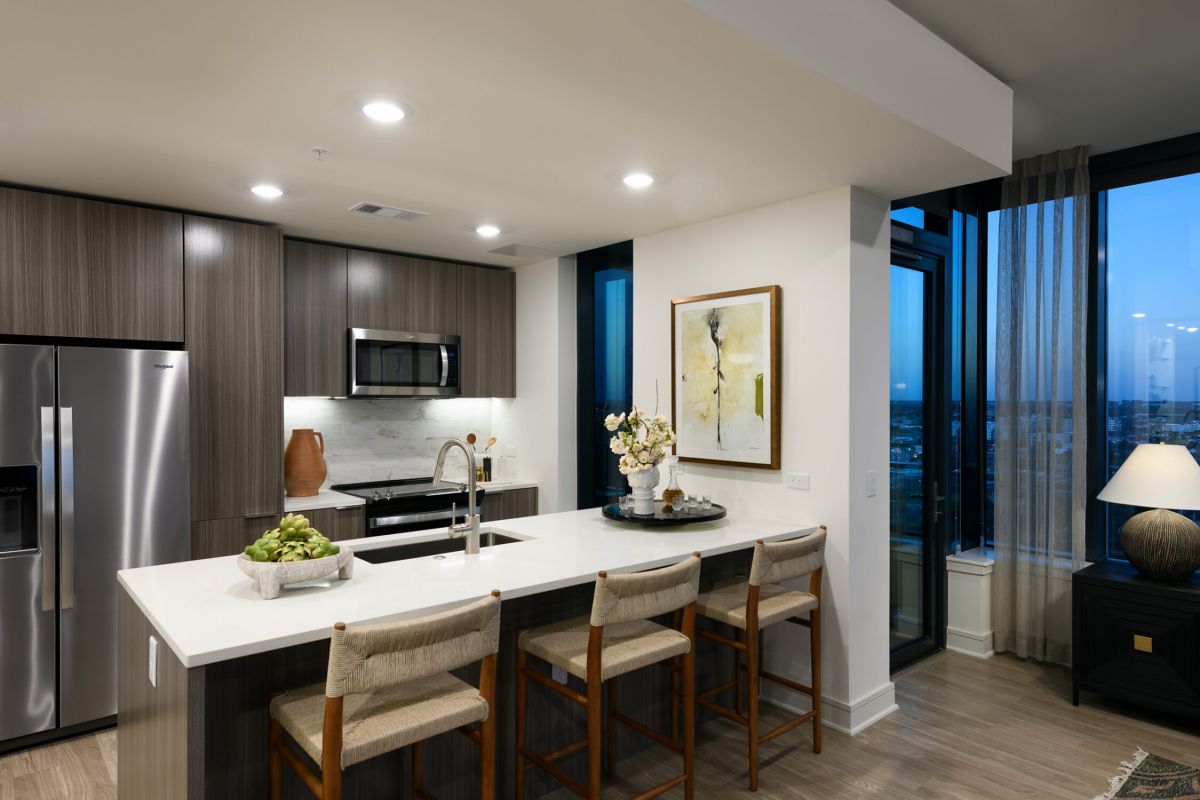 Modern kitchen at Hanover with island, three barstools, stainless appliances, and large windows overlooking Buffalo Bayou at dusk.