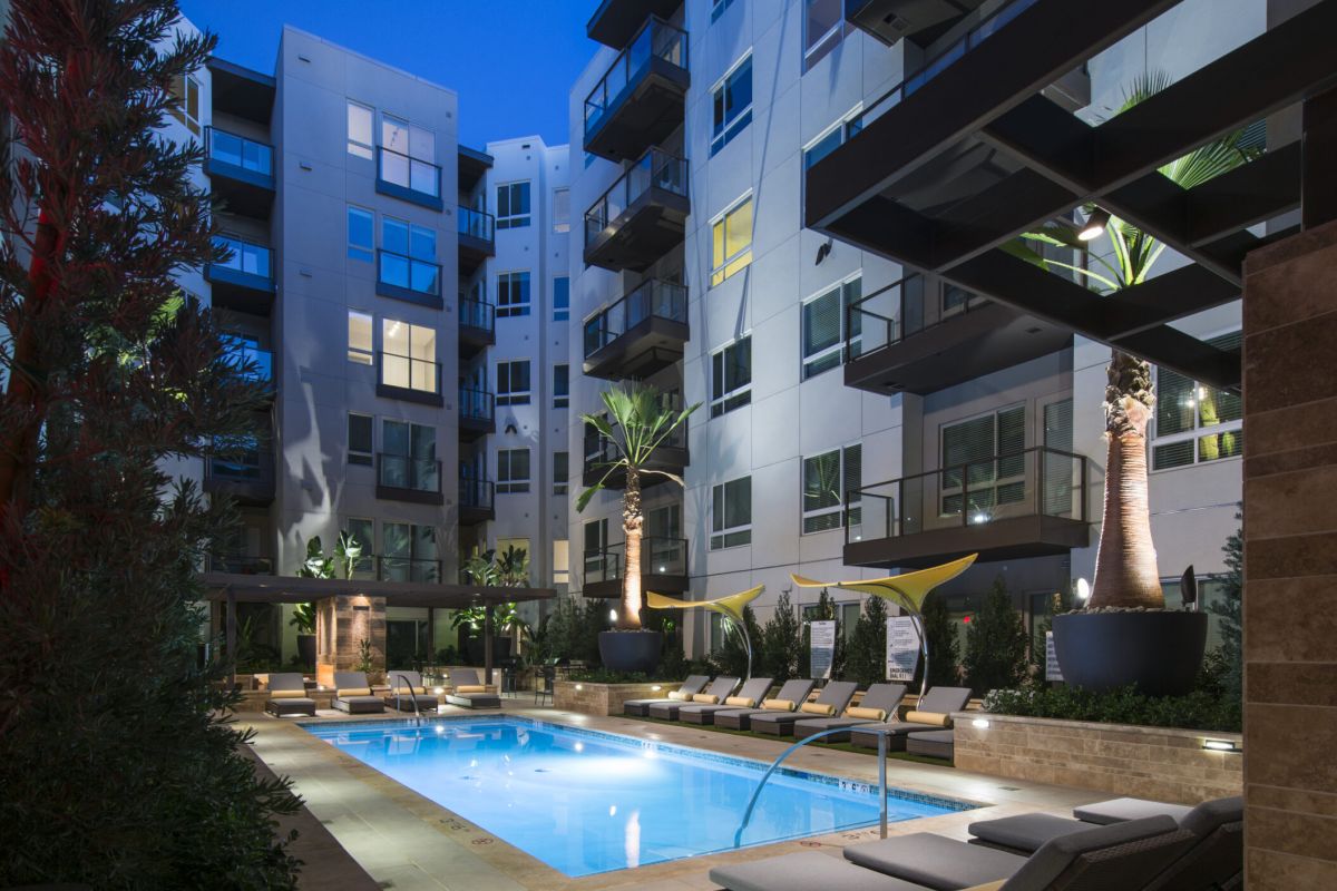 Modern apartment courtyard at night with a lit swimming pool, lounge chairs, and tall palm trees.