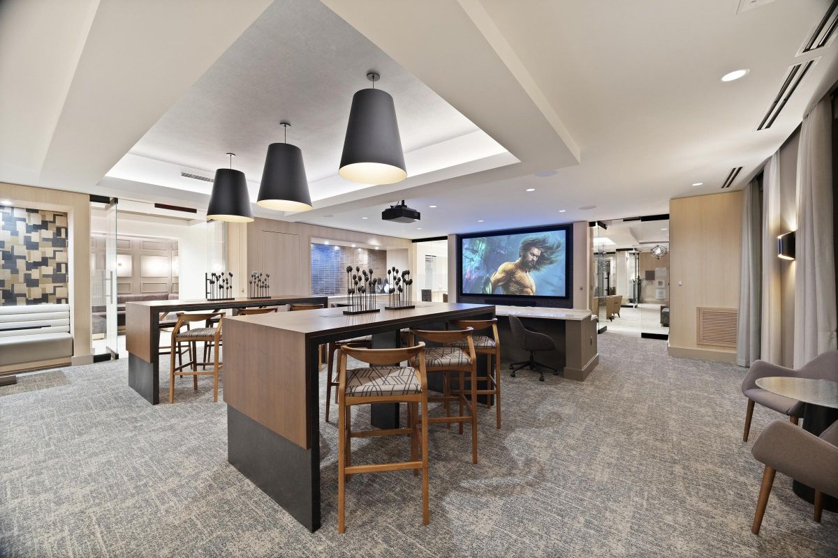 Modern conference room with a large screen, wooden tables, chairs, and black pendant lights.