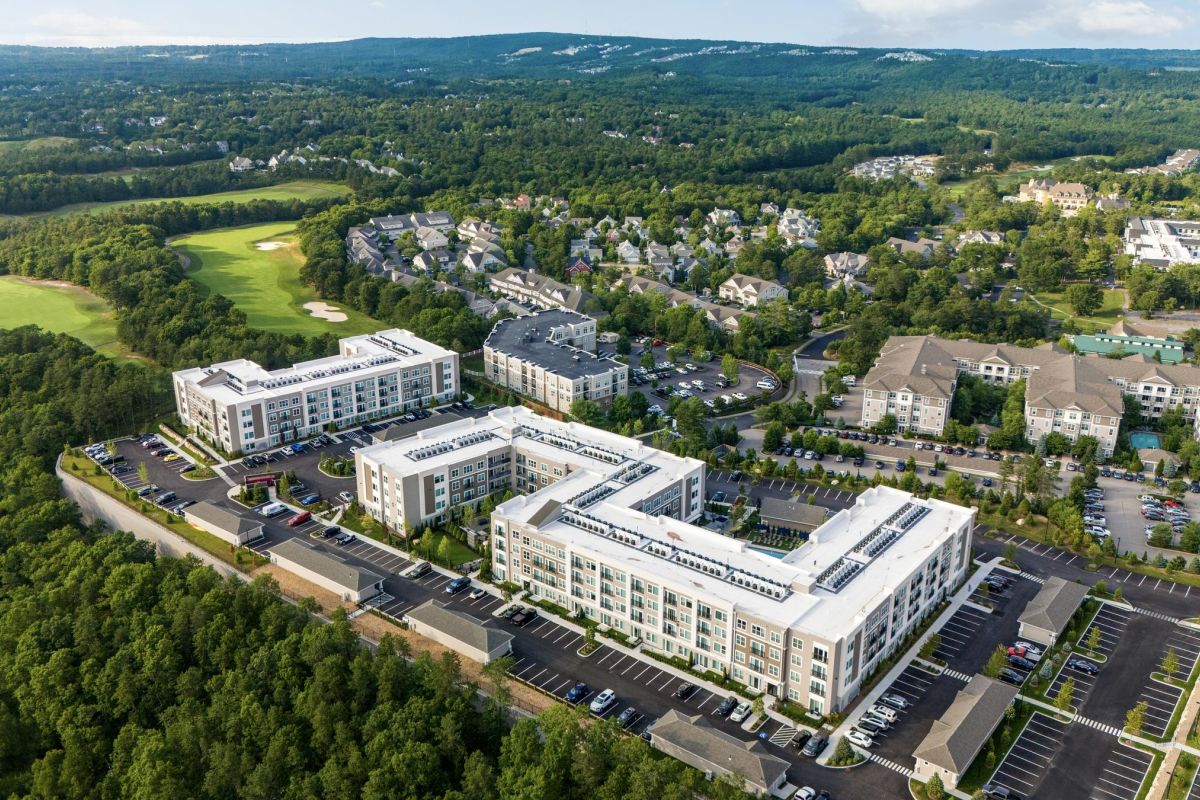 Aerial view of a residential complex with surrounding greenery, roads, and distant hills under a blue sky.