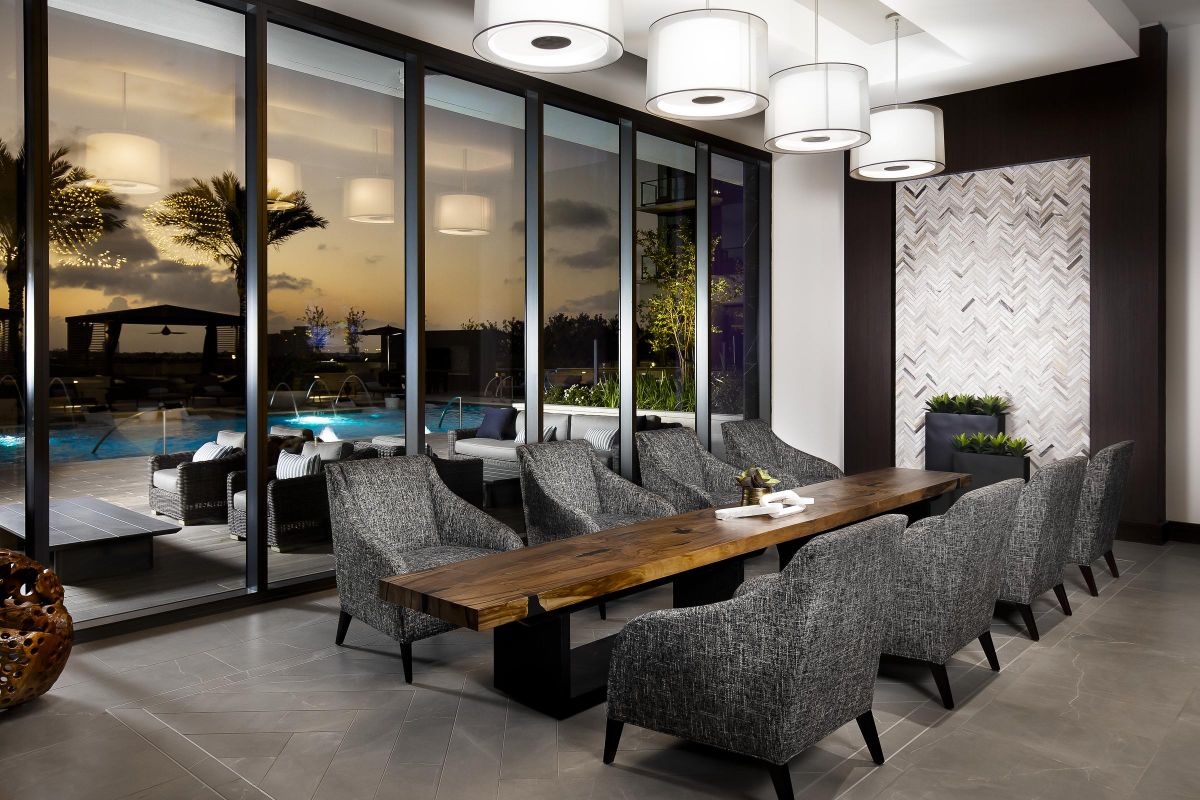 Modern dining room with a long wooden table, gray chairs, and large windows showing a pool and sunset view.