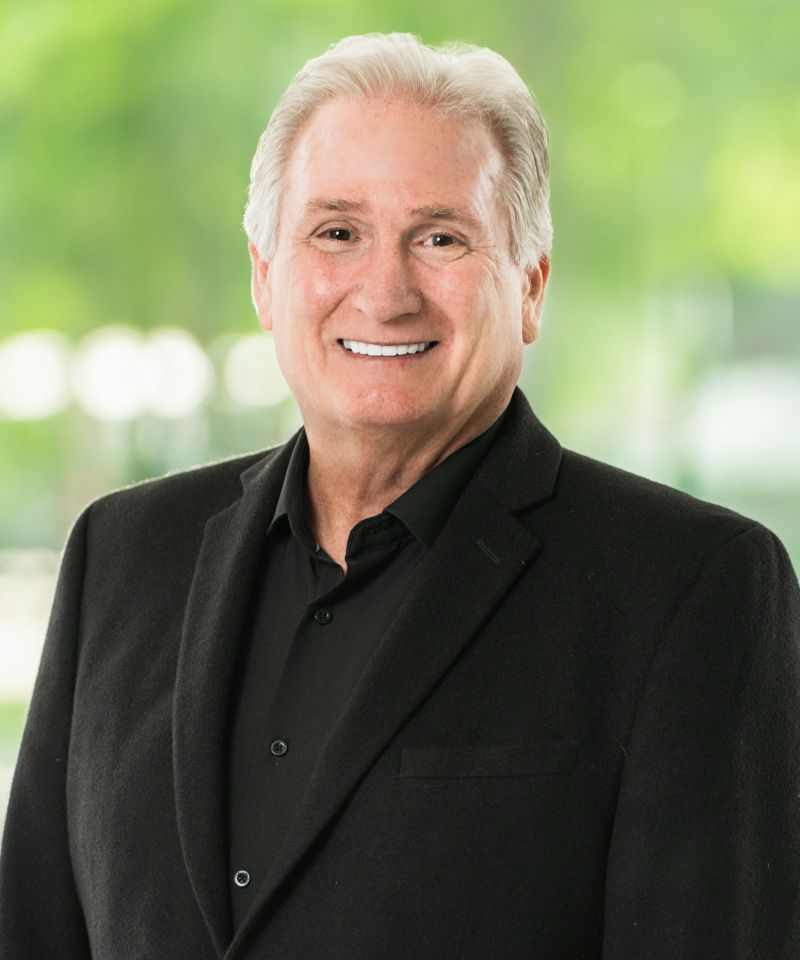 Smiling older man with gray hair wearing a black suit jacket and shirt, standing outdoors with greenery behind.