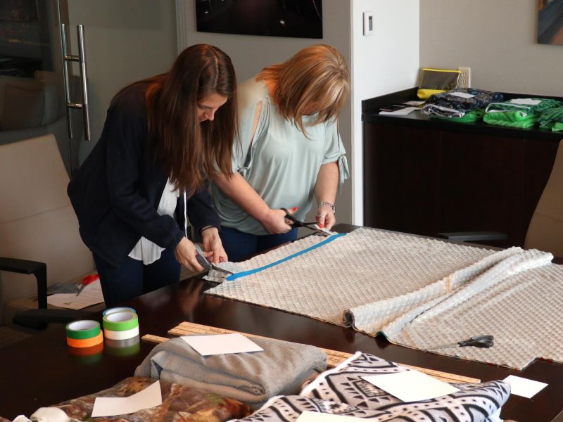 Two women measure and cut fabric on a table, surrounded by supplies and fabric pieces.