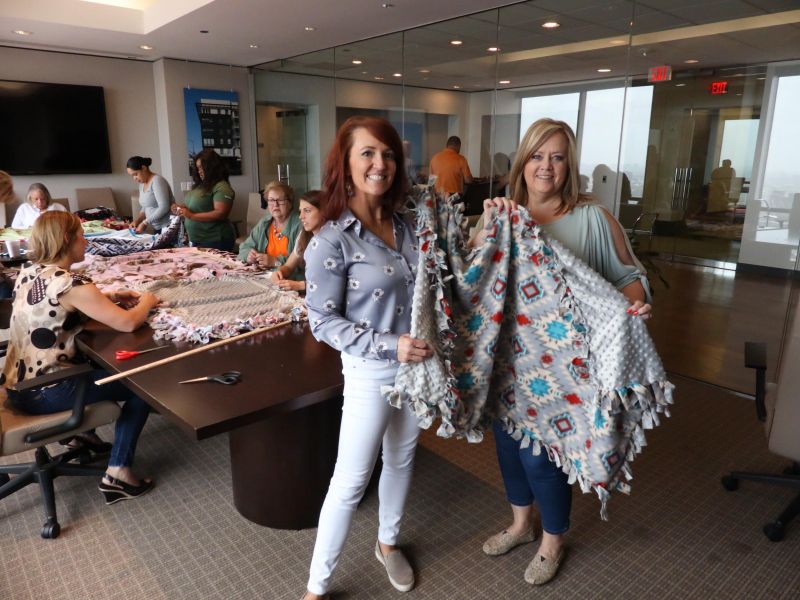 Two women smile and hold up knotted blankets in an office where others are making more blankets at tables.