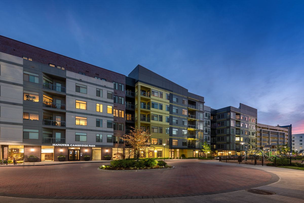 Modern apartment complex at dusk with lit windows and a circular driveway in front.