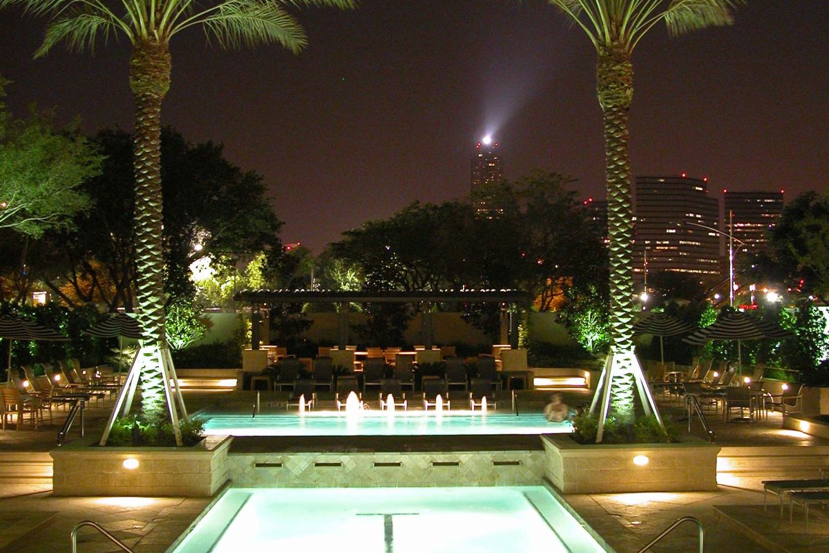 Night view of a lit swimming pool with fountains, palm trees, and city buildings in the background.