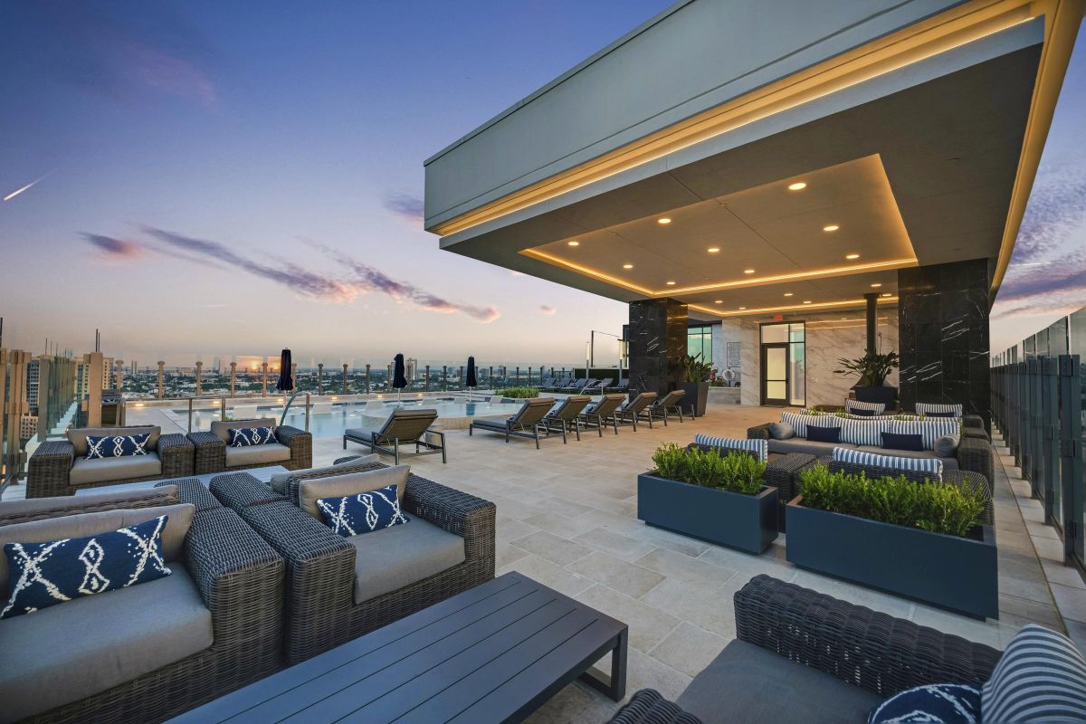 Rooftop lounge with modern seating, tables, and plants, overlooking a city skyline at sunset.
