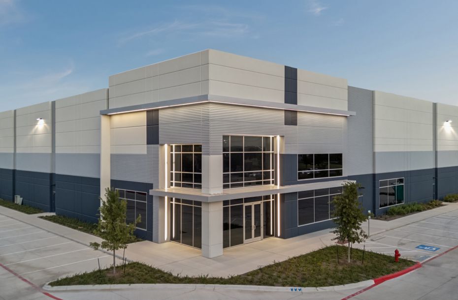 Modern industrial warehouse building with large windows, surrounded by pavement and a few small trees.