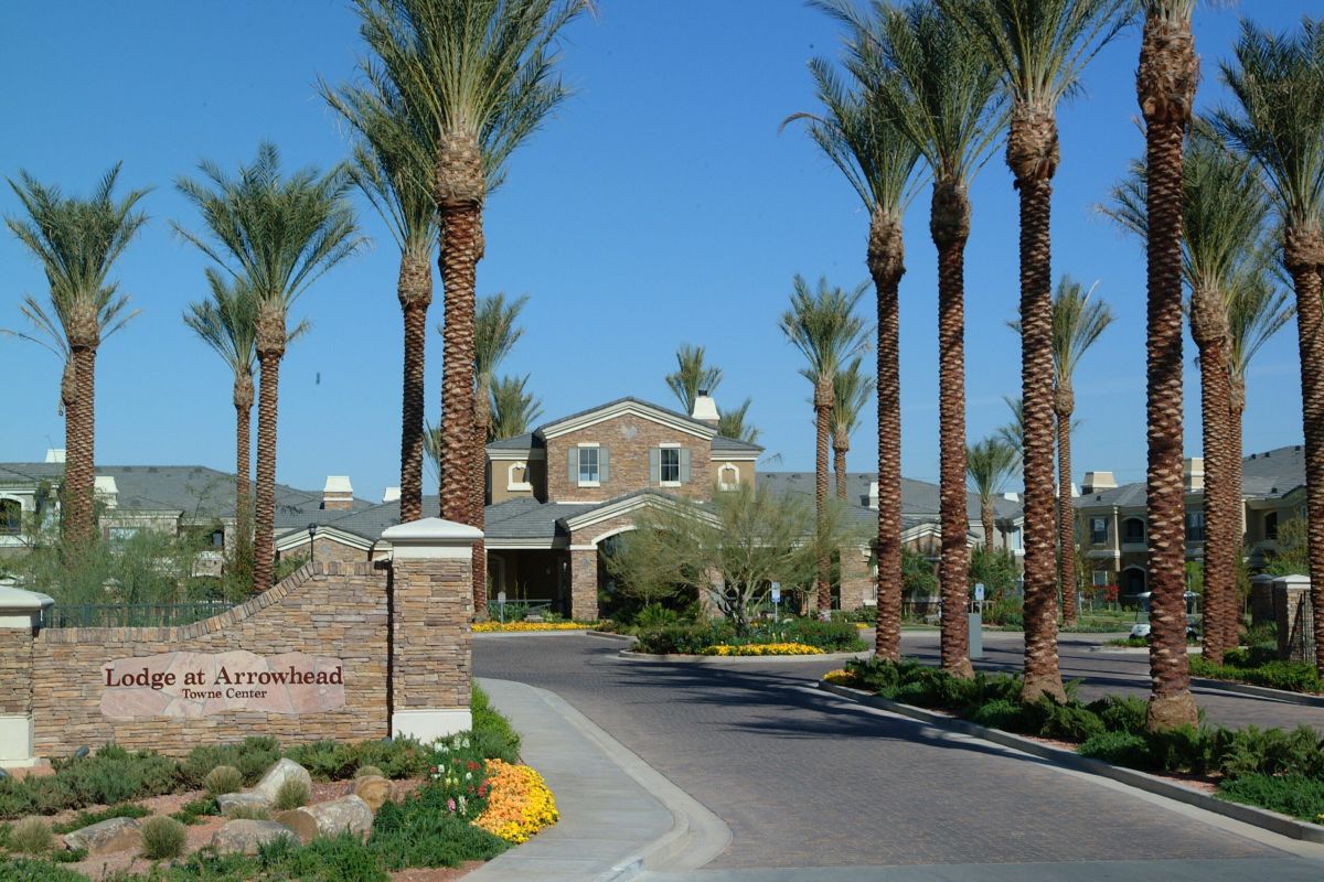 Entrance of Lodge at Arrowhead, lined with tall palm trees and landscaped flower beds under a clear blue sky.