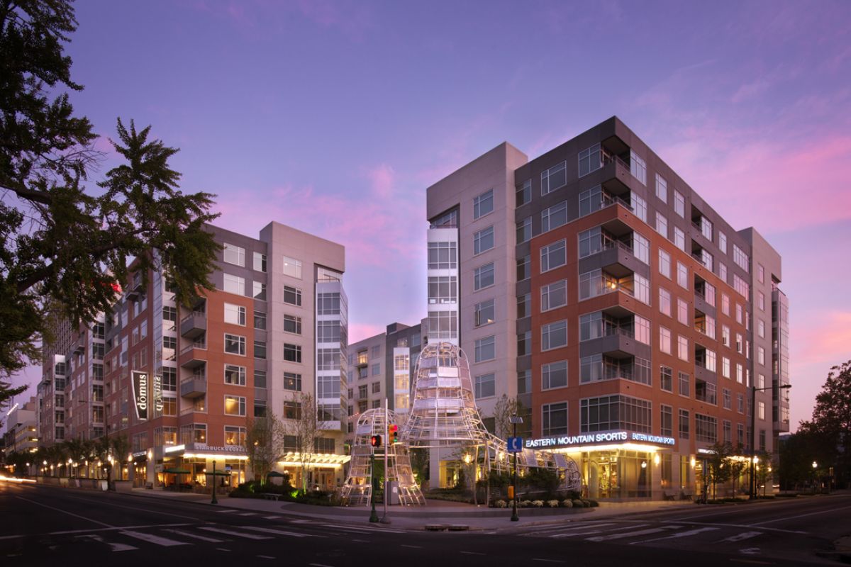 Modern apartment buildings at sunset with streetlights, shops, and a prominent white sculptural structure in front.
