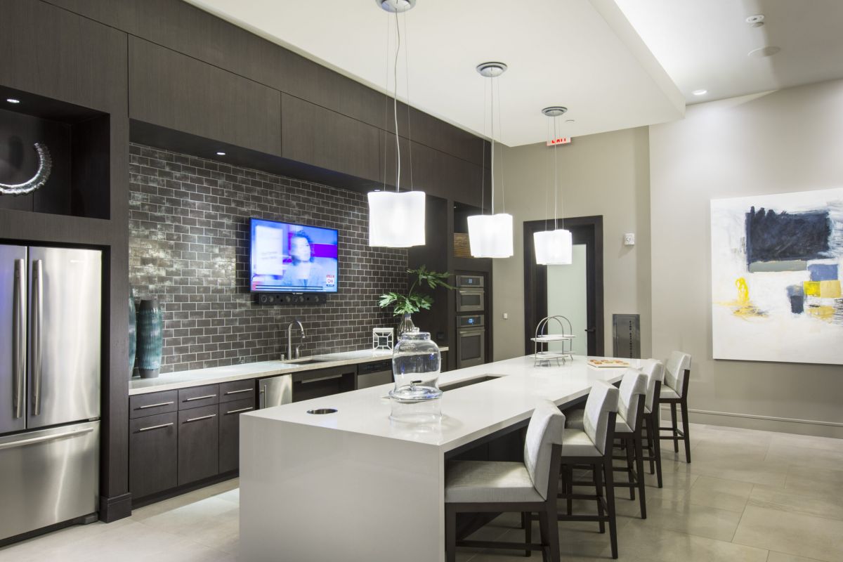 Modern kitchen with dark cabinets, a large white island, bar stools, pendant lights, and a TV on the wall.