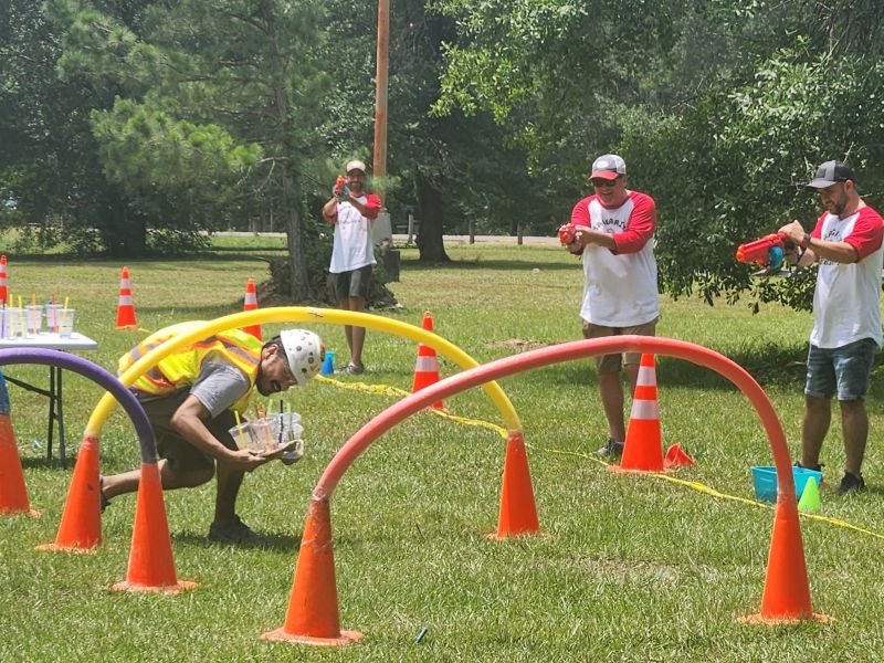 Adults in helmets and red shirts play a water gun game, aiming at cups on a tray outside on a grassy field.