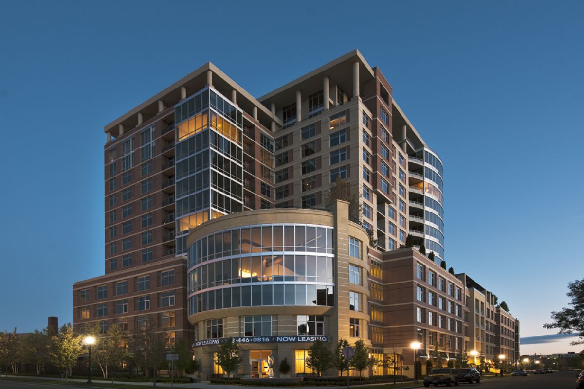 Modern multi-story apartment building with large glass windows, illuminated at dusk, "Now Leasing" banner displayed.