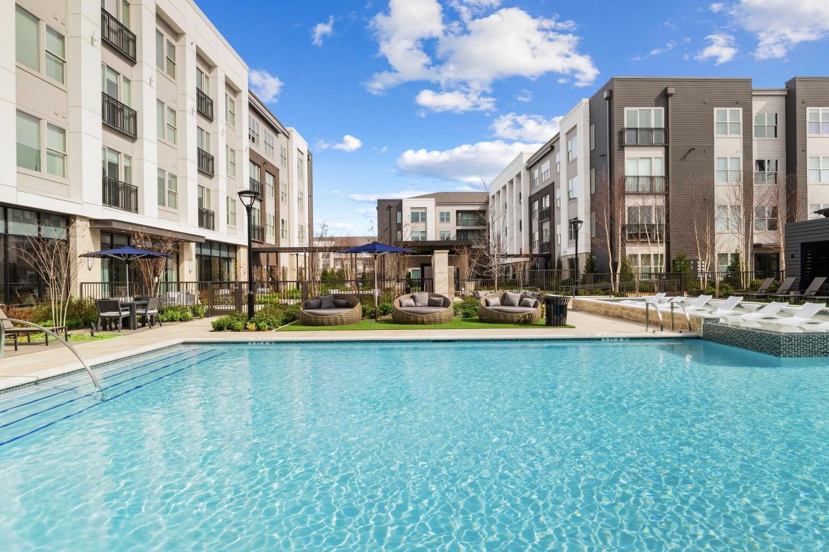 Modern apartments with a swimming pool, lounge chairs, and seating areas under a blue sky with clouds.