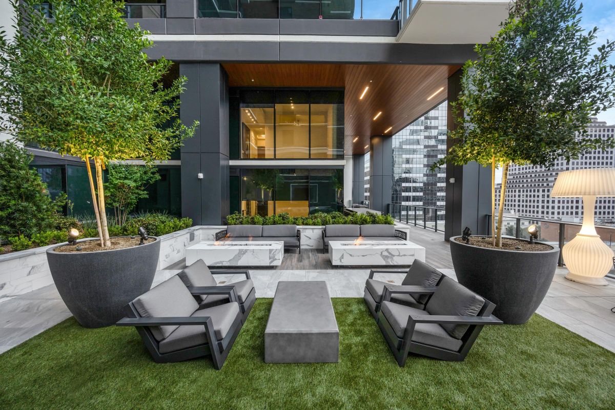Modern outdoor patio with gray chairs, tables, large potted trees, and a cityscape in the background.