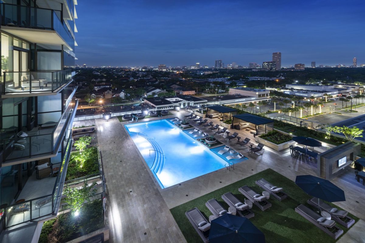 Aerial view of a rooftop pool and lounge area at night, with city skyline in the background.