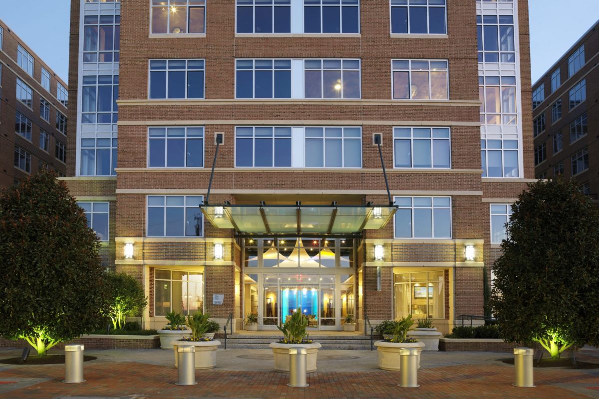 Modern brick apartment building entrance with glass doors, potted plants, and evening lights illuminating the area.
