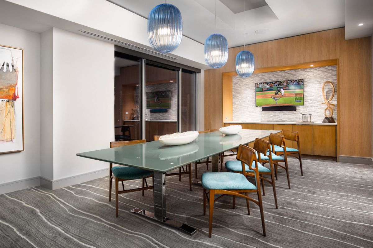 Modern dining room at Hanover Mystic River with glass table, blue chairs, pendant lights, and a TV showing a baseball game.