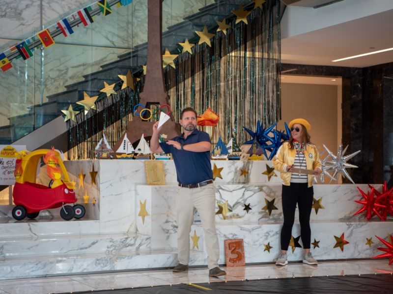 A man throws a paper airplane on a stage decorated with stars; a woman and child watch nearby.