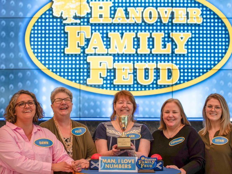 Five women smiling and holding a trophy on a Family Feud game show set, with name tags visible.
