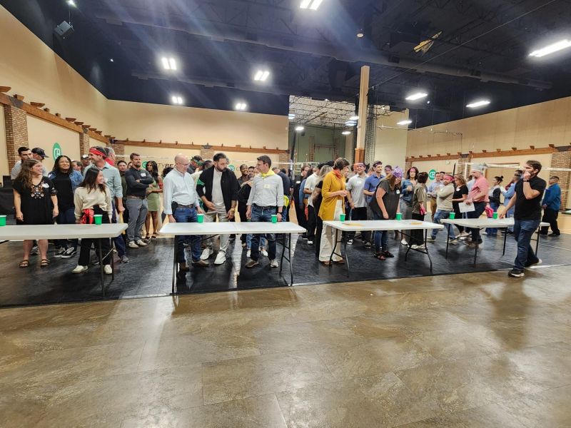 A large group of people gathers around tables indoors, appearing to prepare for a group activity or game.